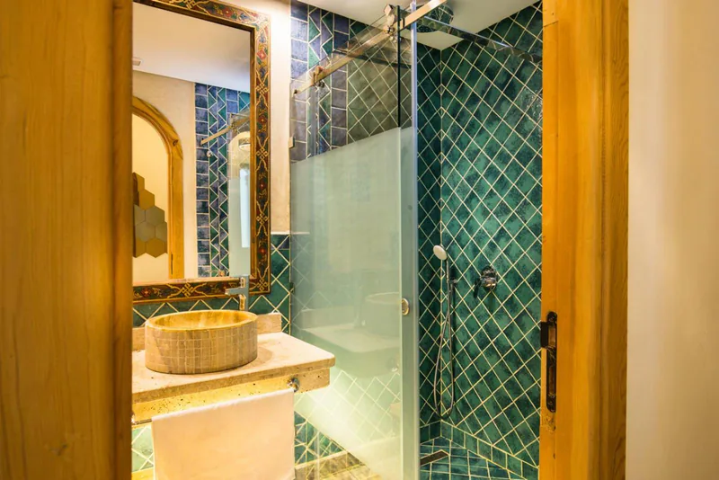 A stone basin under a mirror, beside a glass shower door and an arched wooden door