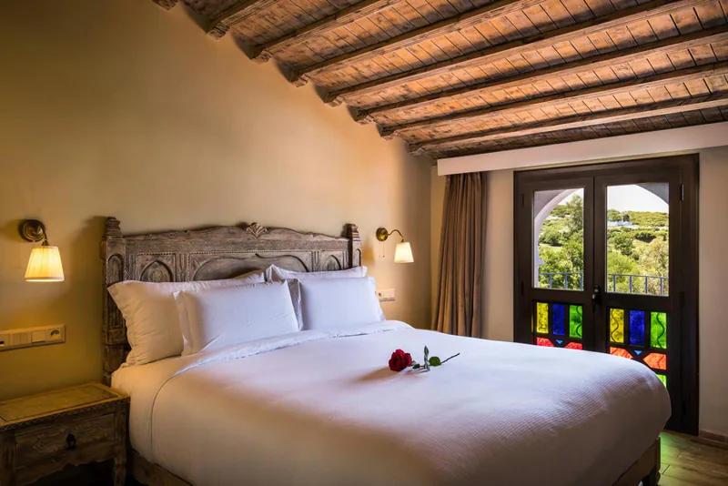 A room with a carved headboard, white linen and a door of coloured glass onto the garden