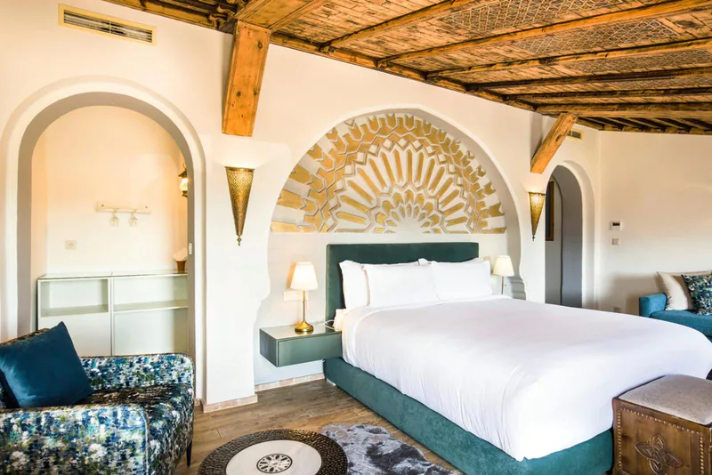 A bed beneath a carved plaster arch, with a teal armchair and a wooden beamed ceiling