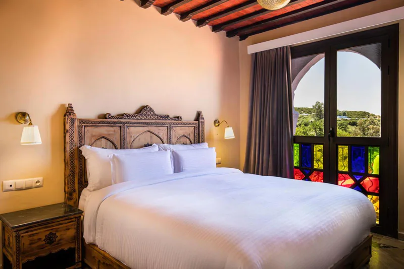 A bed with a carved wooden headboard beside a window of coloured glass
